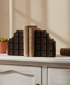 Decorative bookends shaped like chocolate bars on a shelf with books and a plant.