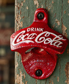 A red cast-iron Coca-Cola bottle opener mounted on a rustic wooden post, featuring the white Coca-Cola script logo and the words 'DRINK Coca-Cola'.