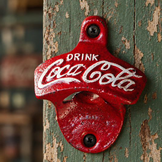 A red cast-iron Coca-Cola bottle opener mounted on a rustic wooden post, featuring the white Coca-Cola script logo and the words 'DRINK Coca-Cola'.