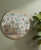 Round decorative wall plate with a botanical and floral pattern, featuring a vintage-style illustration of an armchair and a small table. The plate has a gold-toned rim and is mounted on a light sage green wall next to a green plant.