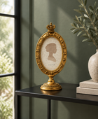 An ornate gold oval picture frame with a crown topper and a silhouette portrait insert, displayed on a black console table in a styled interior setting.