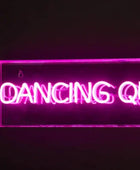 A rectangular neon sign glowing with bright pink/magenta text that reads “DANCING QUEEN,” mounted against a dark wall with a visible power cord. A disco ball is partially visible at the lower left.