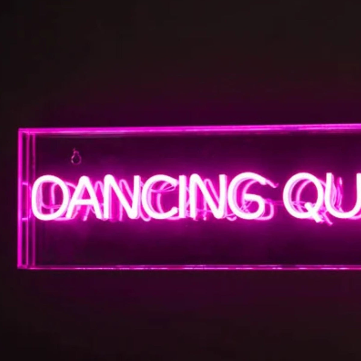 A rectangular neon sign glowing with bright pink/magenta text that reads “DANCING QUEEN,” mounted against a dark wall with a visible power cord. A disco ball is partially visible at the lower left.