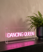 A rectangular clear acrylic LED sign displaying the words 'DANCING QUEEN' in glowing pink neon, placed on a dark wooden surface next to a potted green fern against a textured beige wall.