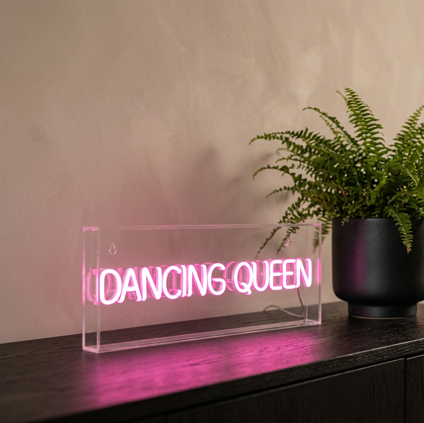 A rectangular clear acrylic LED sign displaying the words 'DANCING QUEEN' in glowing pink neon, placed on a dark wooden surface next to a potted green fern against a textured beige wall.