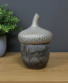 Decorative ceramic acorn-shaped container on a wooden surface with a plant and framed picture in the background.
