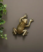 Bronze frog sculpture on a brown wall with a green leafy branch.