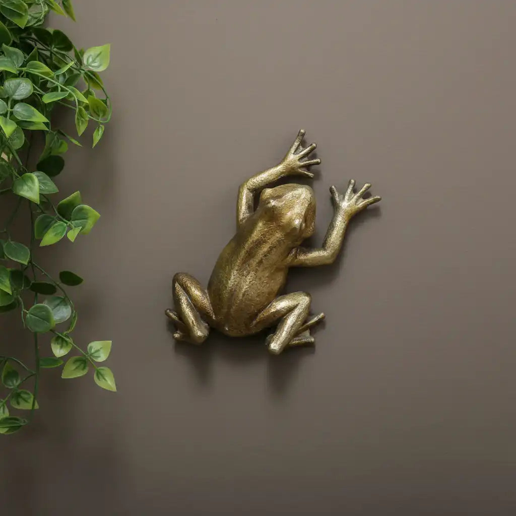 Bronze frog sculpture on a brown wall with a green leafy branch.