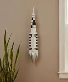 Decorative wall art resembling a rocket on a beige wall with a plant and window in the foreground.