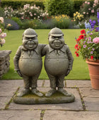 Two stone statues of overweight men in a garden setting with flowers and plants.