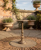 Decorative garden scene with ornate table, urns, and plants on a stone patio.