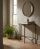 A round wall mirror with a gold-toned frame hangs above a narrow gold console table in a softly lit room with light wood flooring and a potted fern nearby.