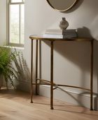 A gold-finished metal console table with a glass top, styled with books and a small vase on a beige wall in a sunlit room with a window and a potted fern.