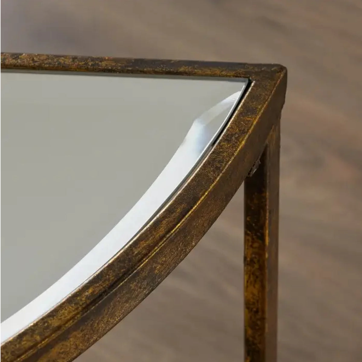Close-up of a corner of a mirrored table with a distressed gold and bronze metal frame, set against a blurred wood floor background.