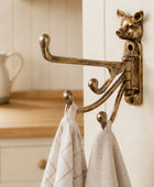 An ornate brass pig-shaped wall hook rack mounted on a white paneled door, holding two beige towels in a kitchen setting.
