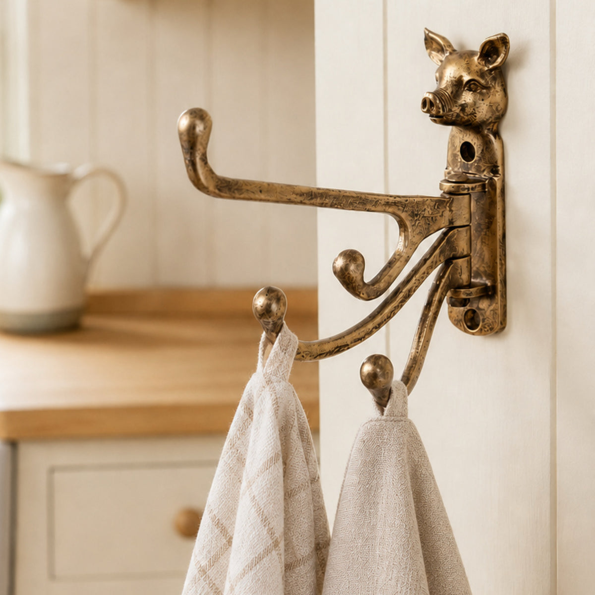 An ornate brass pig-shaped wall hook rack mounted on a white paneled door, holding two beige towels in a kitchen setting.
