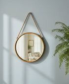 Round mirror with a gold frame hanging on a gray wall next to a potted fern.