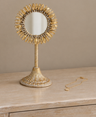 A decorative gold vanity mirror with a sunburst frame and pedestal stand sits on a light wood surface, with a matching gold necklace placed to the right against a warm beige background.