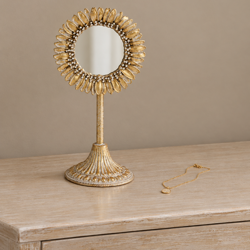 A decorative gold vanity mirror with a sunburst frame and pedestal stand sits on a light wood surface, with a matching gold necklace placed to the right against a warm beige background.