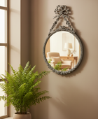 Decorative mirror with floral design on a beige wall next to a potted plant.