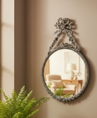 Decorative wall mirror with floral design on a beige wall, next to a window and plant.