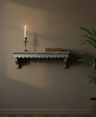 Wall shelf with candle and books against a plain wall, next to a potted plant.