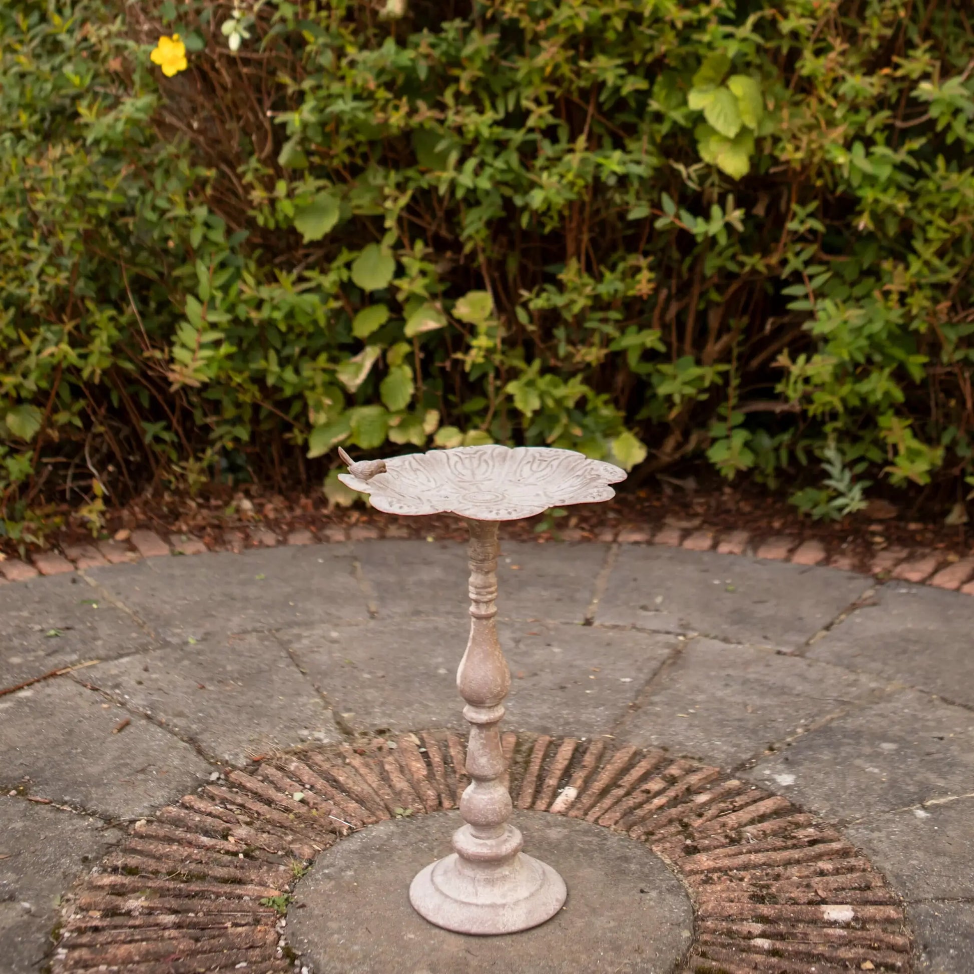 Decorative metal bird bath on a stone patio with greenery in the background