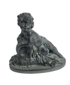 A dark gray resin figurine of a classical nude cherub or putto reclining on a decorative base with draped fabric and fruit details, set against a plain white background.