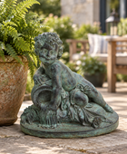 A green patina bronze garden statue of a reclining cherub holding a basket, displayed on a stone patio next to a potted fern.