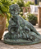 A round outdoor garden statue of a cherub with a green verdigris patina, shown on a stone patio next to a potted fern.
