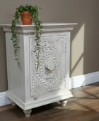 Decorative white cabinet with intricate carvings on a wooden floor against a beige wall.