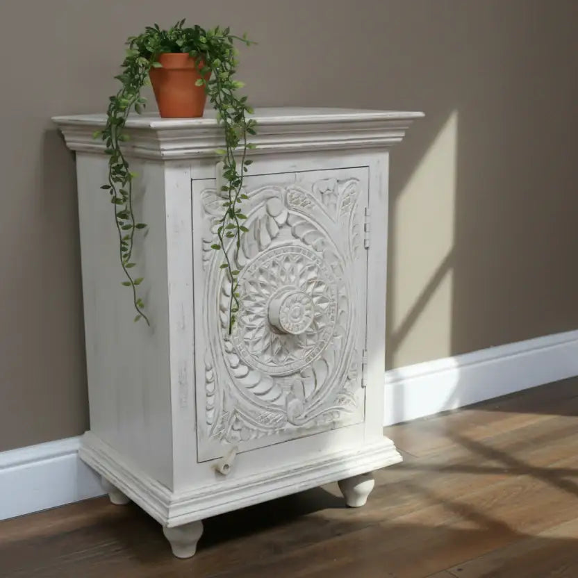 Decorative white cabinet with intricate carvings on a wooden floor against a beige wall.