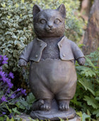 Statue of a cat in a coat and overalls standing among plants and flowers.