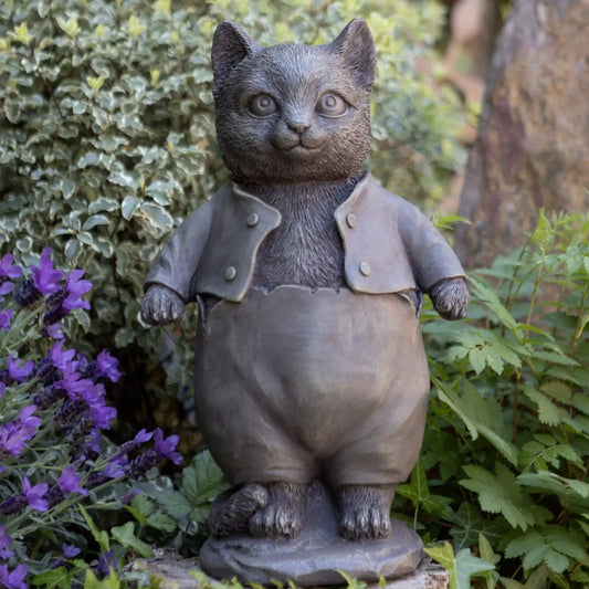 Statue of a cat in a coat and overalls standing among plants and flowers.