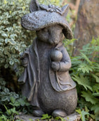 Statue of a rodent wearing a hat and coat, holding a mushroom, in a garden setting.