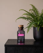 A small dark glass bottle with a cork stopper and a glowing pink neon-style 'dream' sign inside, placed on top of a colorful book or box in an indoor setting.