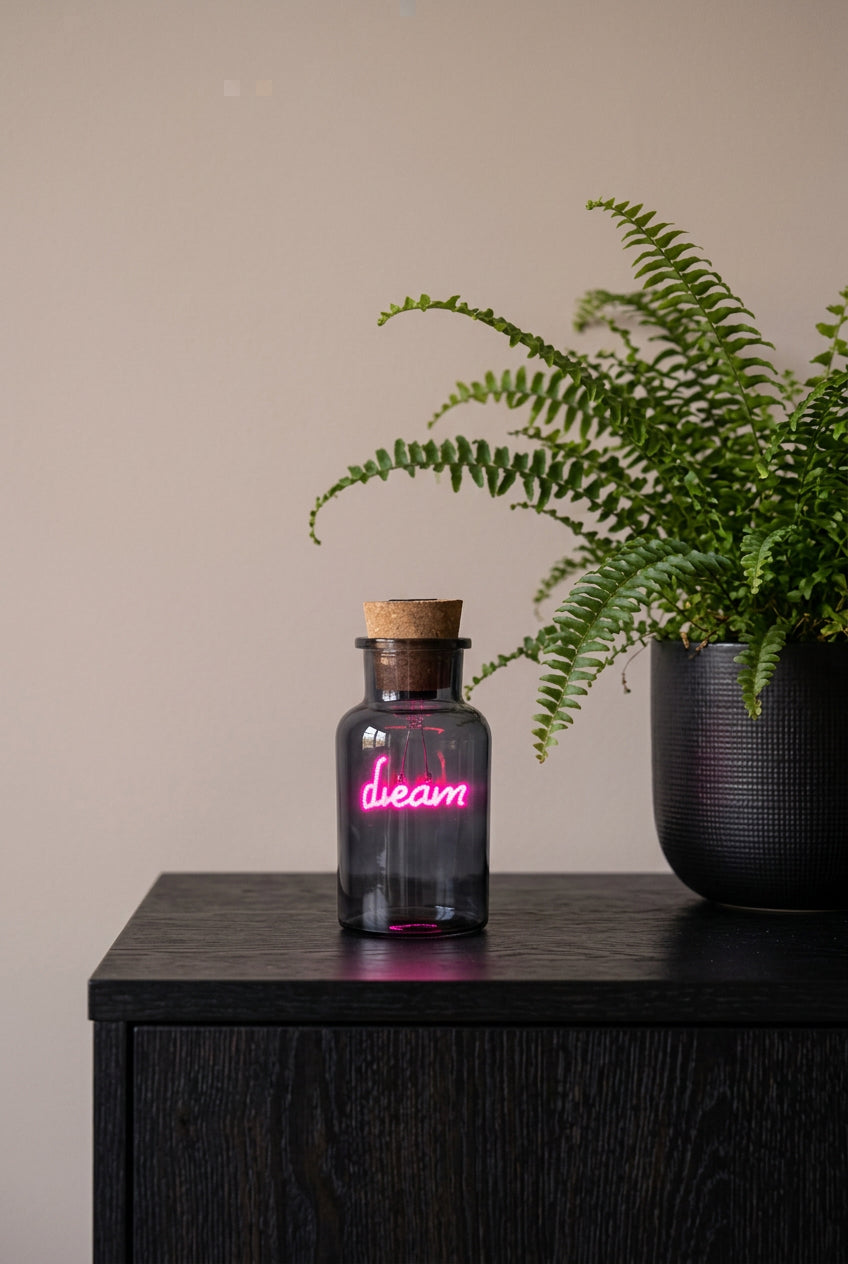 A small dark glass bottle with a cork stopper and a glowing pink neon-style 'dream' sign inside, placed on top of a colorful book or box in an indoor setting.