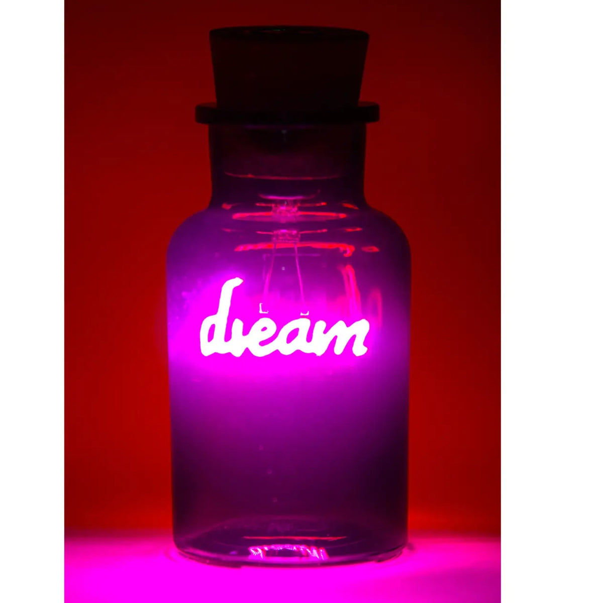 A clear glass bottle with a dark stopper, glowing with a bright pink and purple neon light that spells out the word 'dream' in white script, set against a dark red background.