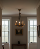 A rustic multi-light chandelier with candle-style bulbs hangs in the center of a hallway with herringbone wood floors, white walls, and two tall windows.
