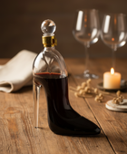 Bottle shaped like a high-heeled shoe with red liquid on a wooden surface, surrounded by wine glasses and a candle.