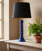 A table lamp with a glossy cobalt blue spiral base and a black drum shade, styled on a light wood table next to a potted fern.