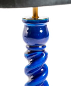 Close-up of a glossy cobalt blue ceramic lamp base with a spiral twist design, featuring a gold-toned metal socket and a dark fabric lampshade at the top, set against a plain white background.