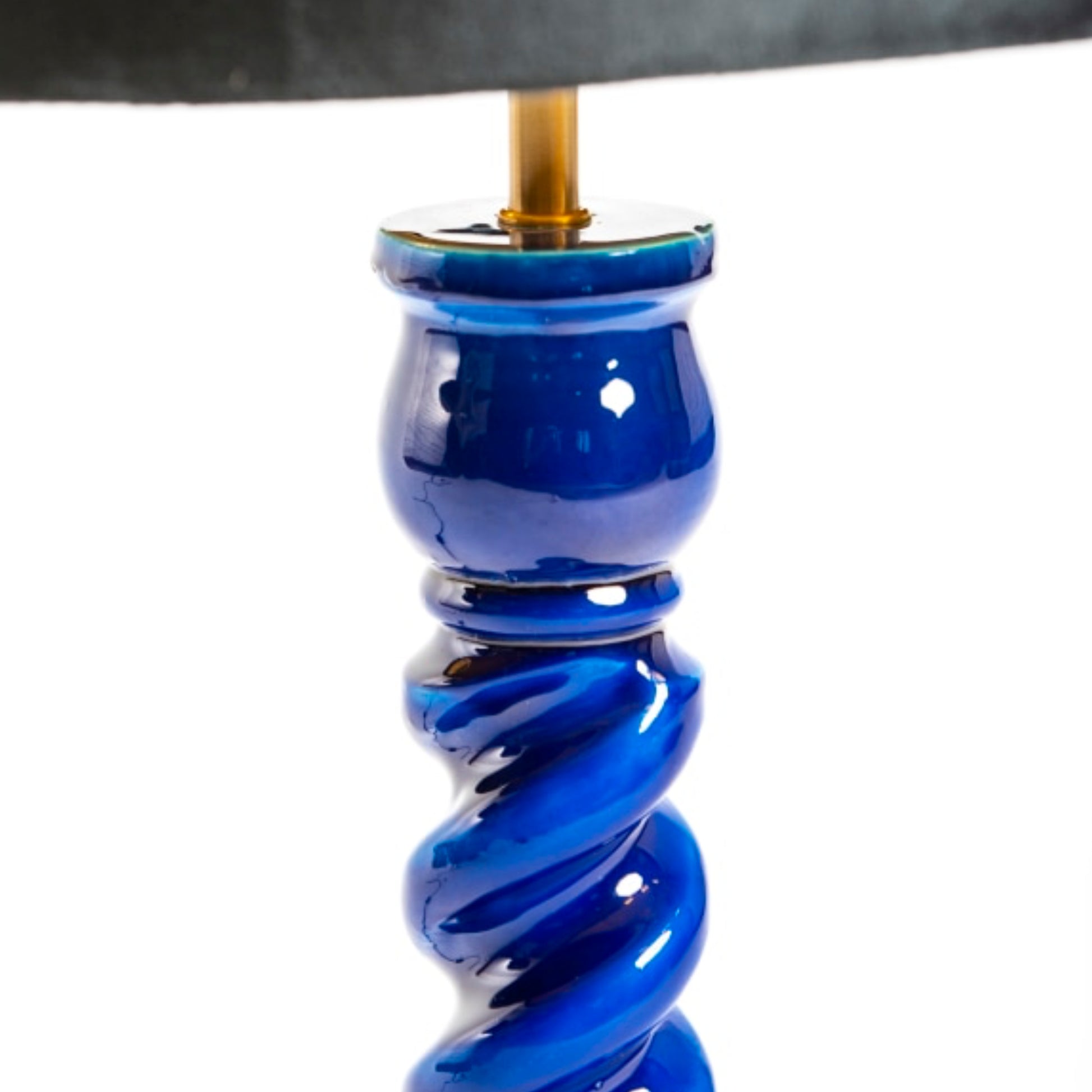 Close-up of a glossy cobalt blue ceramic lamp base with a spiral twist design, featuring a gold-toned metal socket and a dark fabric lampshade at the top, set against a plain white background.