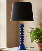 A table lamp with a black fabric drum shade and a glossy cobalt blue spiral pedestal base, shown on a tabletop next to a potted fern in a real interior setting.