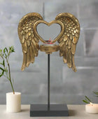 Decorative item with gold angel wings and a heart-shaped design on a stand, with candles and plants in the background.