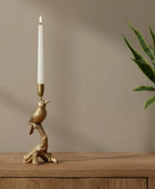 Gold bird-shaped candle holder with a white candle on a wooden surface next to a plant.