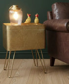 Gold textured side table with a lamp and decorative items next to a brown leather armchair.