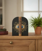 Decorative bookend with eye design on a wooden surface with books and a plant in the background.