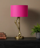 Table lamp with pink shade and gold base on a dark surface against a brown wall.
