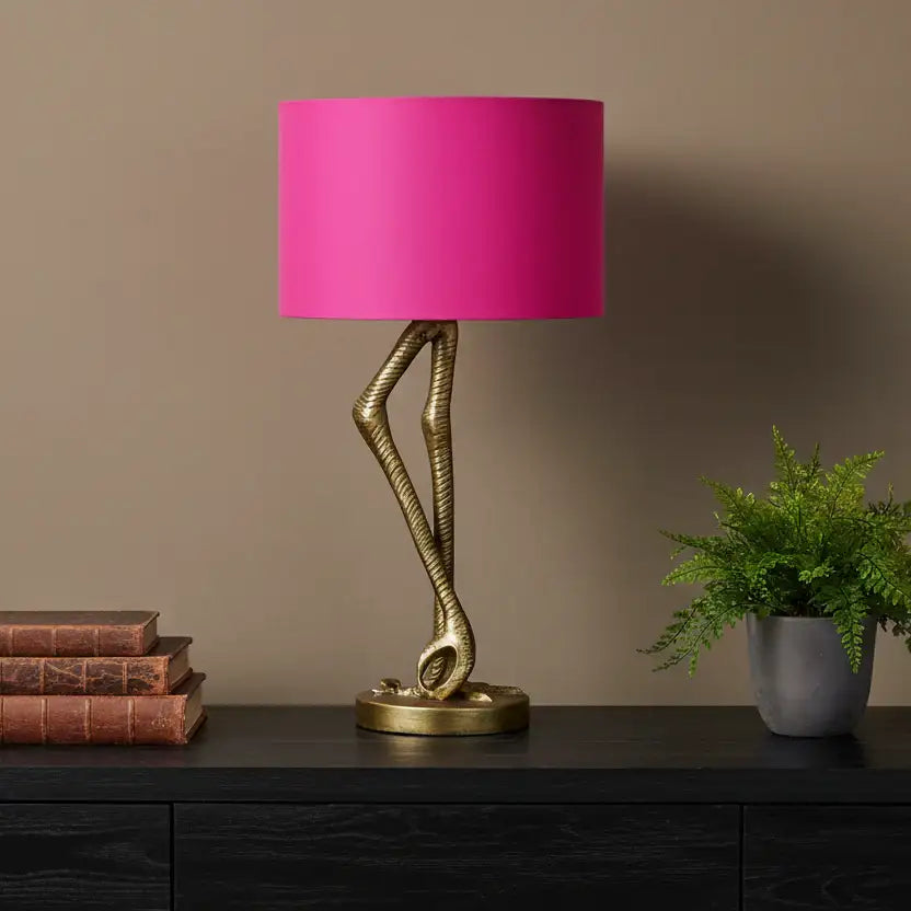 Table lamp with pink shade and gold base on a dark surface against a brown wall.